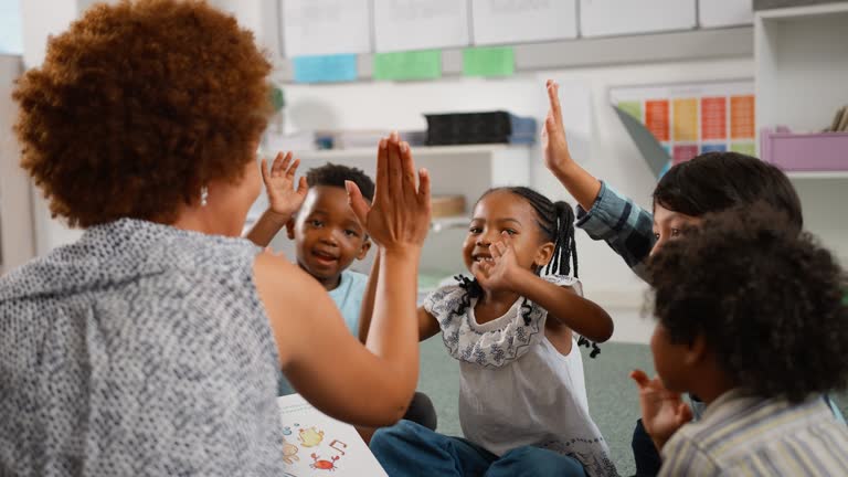 Female Teacher Reads And Gives High Fives To Multi-Cultural Elementary School Pupils In Class