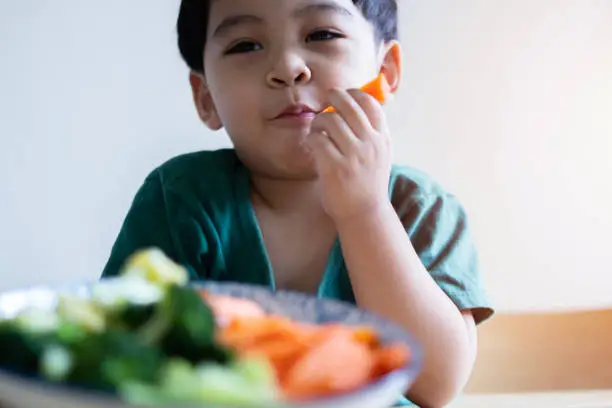 Photo of Portrait of cheerful asian boy eating salad with joy. He is looking aside with curiosity and smiling.