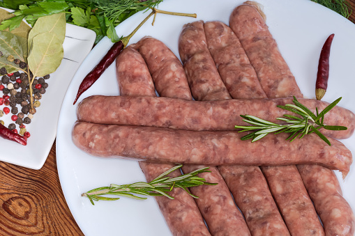 Raw thin long sausages for grilling in the natural casing on a dish among the some spices and fresh greens on the old rustic table, fragment close-up