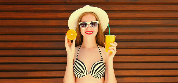 Summer portrait of happy smiling young woman drinking fresh juice with slice of orange fruits wearing straw hat, bra
