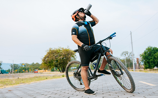 Chubby cyclist on his bike drinking water, Thirsty cyclist on his bike drinking water outside. Tired cyclist drinking water outdoors