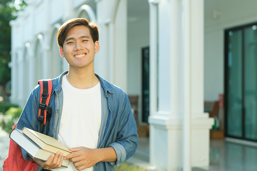 Young smart male collage student in casual clothings and backpack smiling, standing outdoor with campus building in the background and holding books while looking at camera ready for study. Education concept.
