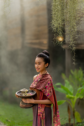 Half body shot of a young and beautiful Lao women wearing traditional Laos costume outside a lush tropical park holding a traditional offering tray with pedestal