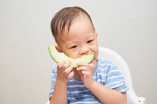 Portrait of Cute Smiling Asian 18 months, 1 year old toddler baby boy child eating melon, Little kid sitting in high chair eating fruit, Healthy snack and Self feeding concept