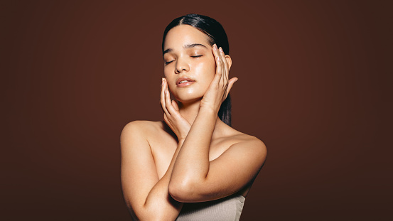 Beautiful young woman touching her flawless skin in a studio. Gorgeous Hispanic woman stands with her eyes closed in a studio, showing off her smooth and soft skin; the results of her skincare routine.