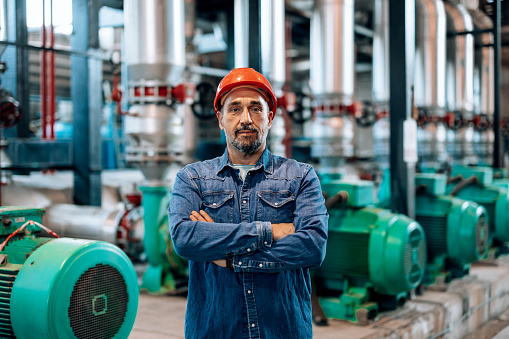 A mature Caucasian engineer is standing in a heating plant with his arms crossed and looking at the camera.