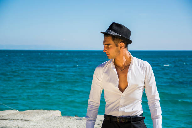 Young handsome man in elegant white shirt and black fedora hat, on beach while looking away. Sea waves on background