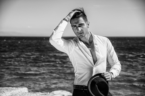 Young handsome man in elegant white shirt and black fedora hat, on beach while looking away. Sea waves on background