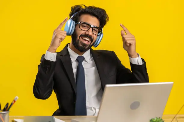 Photo of Indian young business man in headphones listening favorite disco music, working on laptop at office