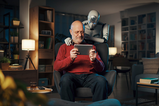 Happy senior man watching videos on his tablet and interactive humanoid AI robot standing next to him