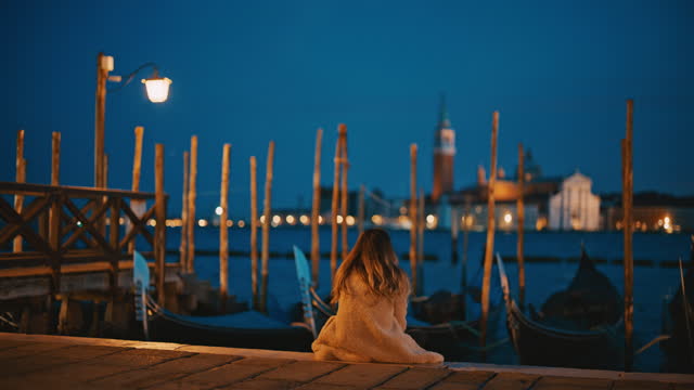 Slow motion of female tourist sitting at jetty. Woman sitting in front of gondolas moored at canal in Venice, Italy. Exploring city at dusk.