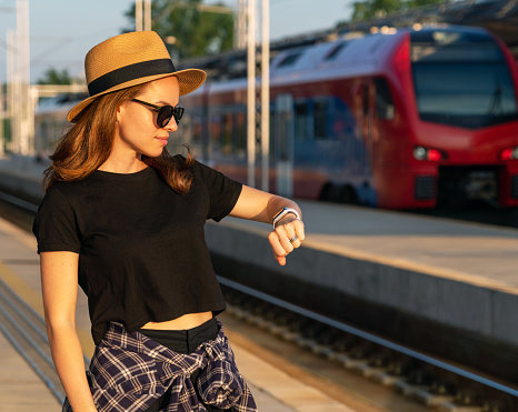 Woman in straw hat and sunglasses standing on railway platform and waiting her train, looking at watch.
