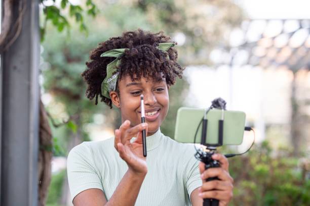 istock Female with curly hair applying makeup while looking at smartphone on selfie stick 1488575292
