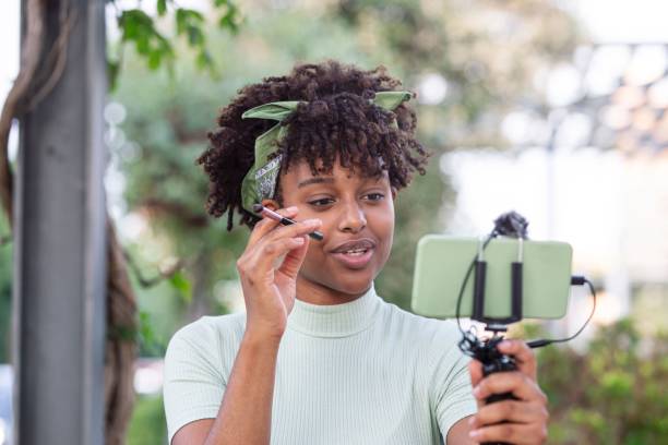 istock Female with curly hair applying makeup while looking at smartphone on selfie stick 1488575296