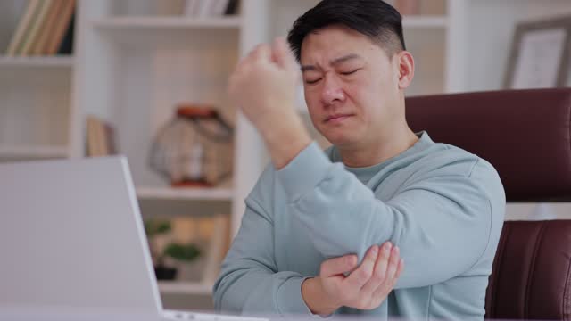 Pain in elbow. Asian man at computer workplace and experiencing pain and discomfort in elbow joint. Arthritis, orthosis, bursitis concept Pain in elbow. Arthritis, orthosis, bursitis concept