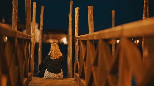 Slow motion of female tourist sitting at jetty. Woman is sitting in front of canal. Exploring city at night.