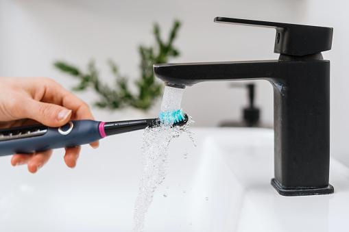 cropped shot of female hand wash electric teeth brush with water stream from black faucet in domestic bathroom, dental care and hygiene concept