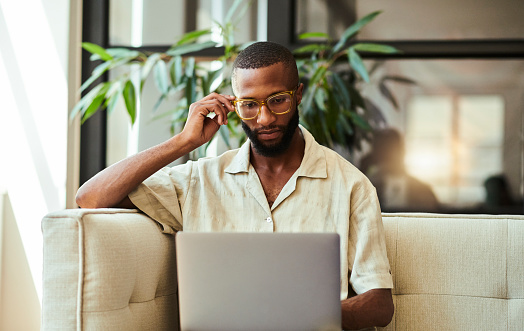 Young black man with a beard sitting on his couch or sofa, casually dressed wearing eye glasses whilst scrolling through the internet using his laptop computer. stock photo