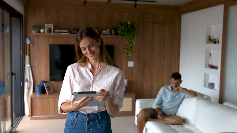 Beautiful woman holding a tablet looking very cheerful daydreaming while partner is sitting at the back on a couch