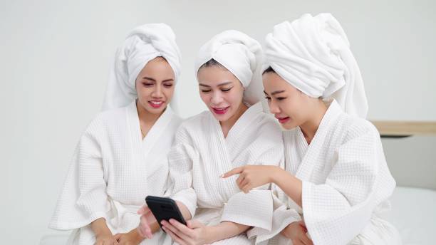 istock Three female friends in bathrobes and towels on head sitting on bed while enjoying using smartphone. Friendship concept 1488591841