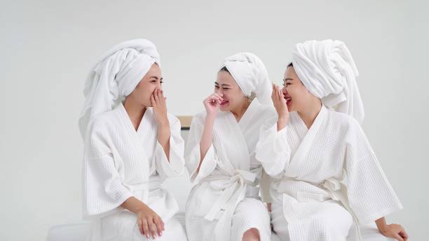バスローブを着て頭にタオルをかぶった3人の若い美しいアジア人女性がおしゃべりを楽しんでいます。ガウンとタオルを着た女性の親友が楽しんでいる - hotel hotel room bed luxury ストックフォトと画像