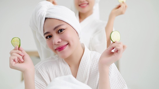 Portrait of beautiful young asian women in bathrobe and towels on heads holding slices of fresh cucumber making refreshing eyes mask. Skin care concept