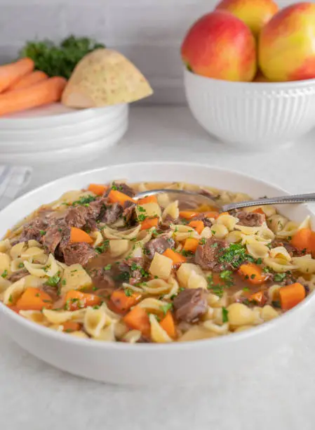 Delicious homemade beef noodles soup with root vegetables. Served hot and ready to eat with spoon on a plate or bowl on white kitchen counter at home. Closeup, front view