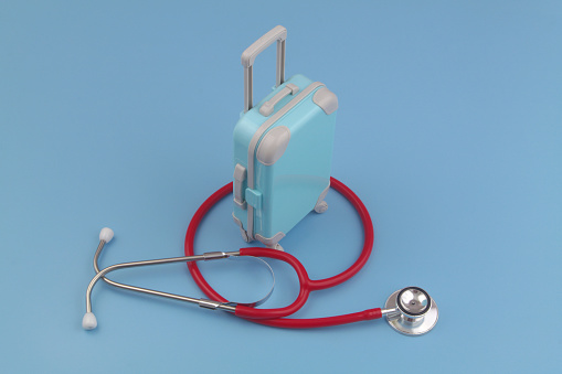 Travel insurance concept. Red stethoscope and travel suitcase on blue background.