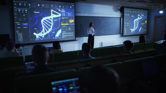 Young University Professor Explaining a DNA Sequencing Methods to a Group of Diverse Multiethnic Students in a Dark Auditorium. Female Teacher Showing a Macro Image of a DNA Structure on a Big Screen
