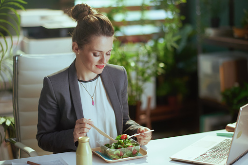 Sustainable workplace. modern middle aged accountant woman in a grey business suit in modern green office with laptop eating salad.