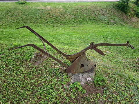 An old rusty plow on a tree stump in the middle of a green lawn on the outskirts of the village.