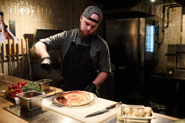 istock Young professional chef in uniform spreading ingredients on pizza flatbread 1488606270