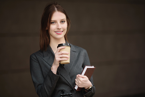 Young businesswoman in a business suit with a notebook and coffee on the street looks into the frame.The concept of a break in students, journalism at work. High quality photo