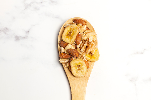 Almonds, walnuts, peanuts and dry banana slices on a wooden spoon and on a white kitchen counter with copy space