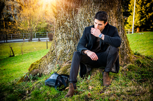 Handsome young man leaning against tree in autumn, wearing black coat