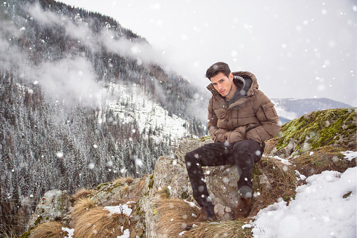 Handsome young man in winter outerwear sitting under snow up the mountain, while looking at camera