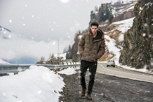Young man walking on roadside under the snow up in the mountains. Snowy mountain on background