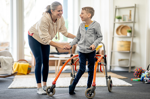 A Mother works with her young son at home on his mobility.  They are both dressed comfortably as the boy sits on his walker and his Mother lends some assistance.