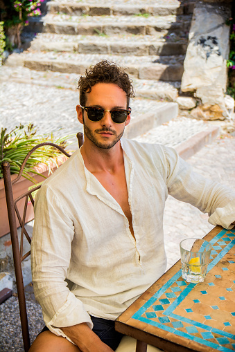 Portrait of stylish good-looking man in sunglasses holding bottle of water at table in cafe