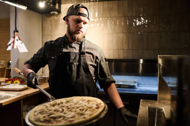 istock Bearded chef in grey uniform and cap standing in front of electric oven 1488614887
