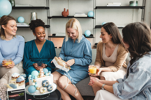 A young beautiful pregnant woman opening presents at a baby shower party. She is sitting on a couch in her beautifully decorated living room surrounded by her best friends. They look excited and happy.