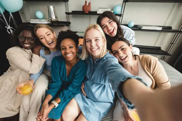 A young Caucasian mother-to-be taking a selfie with her close friends at her baby shower to commemorate the moment. They look happy and cheerful. They are hugging and leaning on to each other showing how close they are.