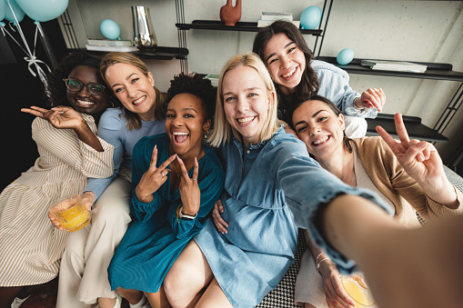 A young Caucasian expectant mother taking a goofy selfie with her diverse friends at her baby shower celebration. The girls are looking at the camera and smiling while posing with a peace sign. They look cheerful and are having a good time together.