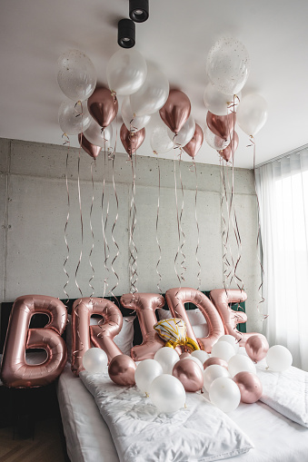Beautiful balloons on a bed in a hotel room spelling the word bride. Beautiful decorations for a bachelorette party. The balloons are white and pink. The room is minimalistic.
