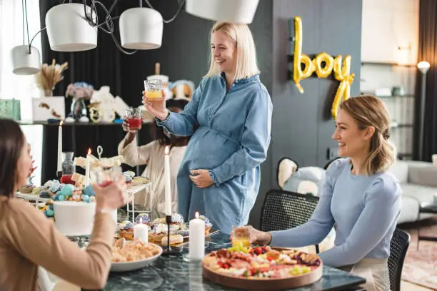 A young Caucasian expectant mother raising a toast at her own baby shower. She is holding her belly and raising her glass while her close friends are sitting next to her. She is giving a speech to thank her friends for planning such a beautiful baby shower.