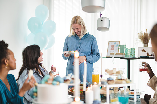 A happy caucasian mother-to-be holding her pregnant belly while hanging out with her friends at the baby shower. They are all laughing and having fun together while celebrating the special occasion. The apartment is full of baby shower decorations and gifts. Everyone looks happy.