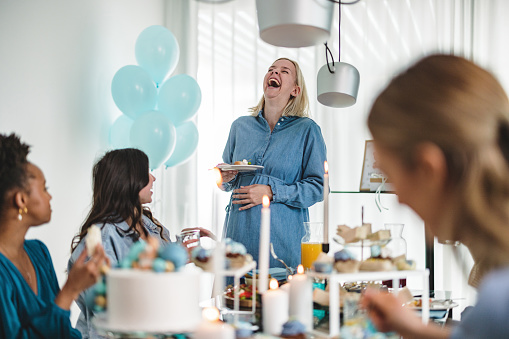 A young Caucasian mother-to-be laughing during a baby shower planned by her best friends. She is having fun while hanging out with the girls. They are joking around while eating delicious food.