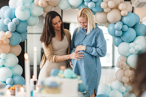 A brunette Caucasian female holding her pregnant best friend's belly at a baby shower. They are standing in an apartment and have a balloon ring behind them. They are smiling and look happy. Beautiful heartfelt moment.