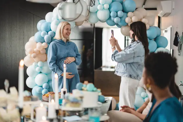 A brunette Caucasian female taking a photo of a young Caucasian expectant mother at her baby shower to commemorate the moment. The rest of the girls are sitting by the table full of delicious food. They are happy to be hanging out and celebrating such a beautiful moment together.