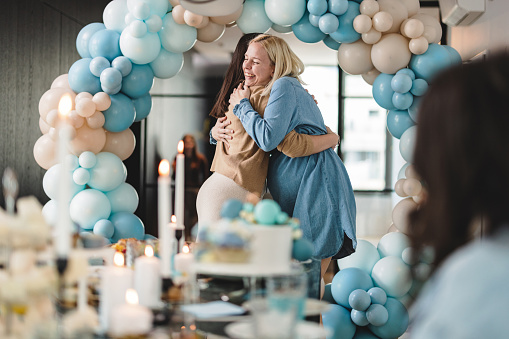 A heartwarming moment of best friends hugging at a baby shower. A beautiful baby shower celebration in an apartment decorated with blue balloons.